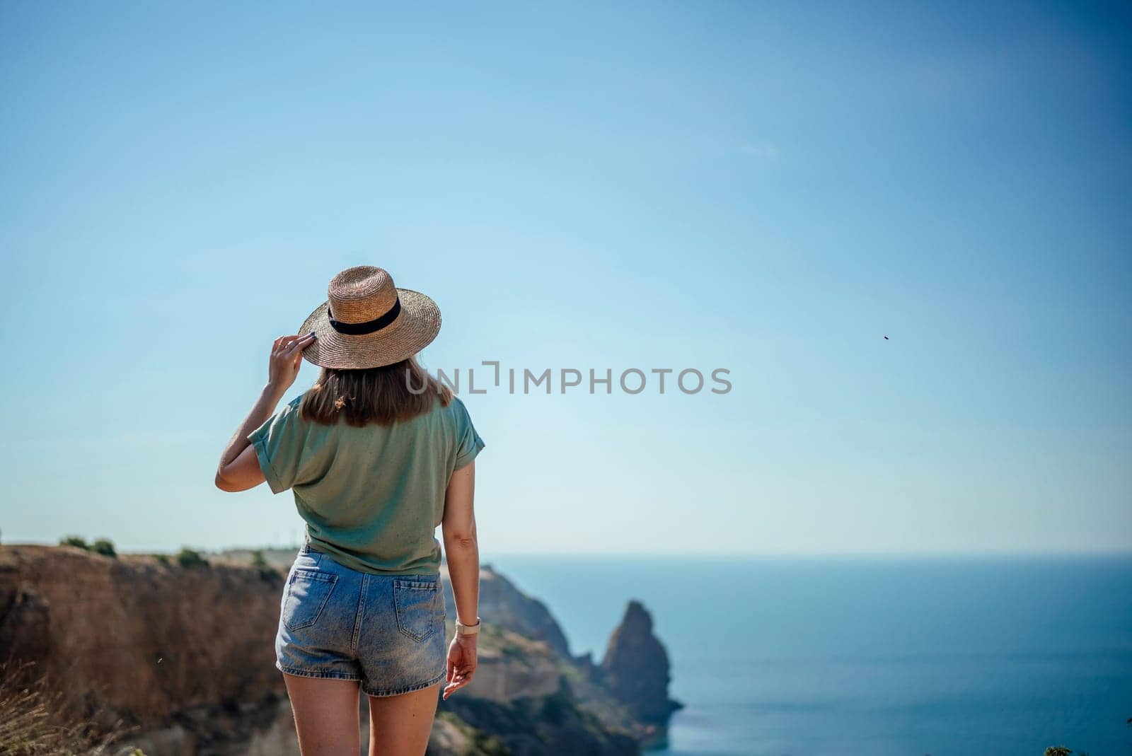 A woman wearing a straw hat stands on a cliff overlooking the ocean. She is wearing blue shorts and a green shirt