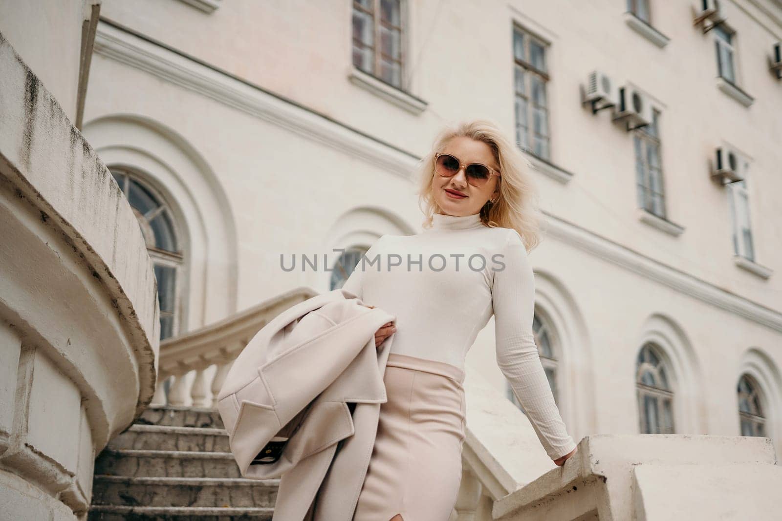 A woman wearing a white shirt and skirt is standing on a staircase in front of a building. She is holding a handbag and she is posing for a photo. Concept of elegance and sophistication