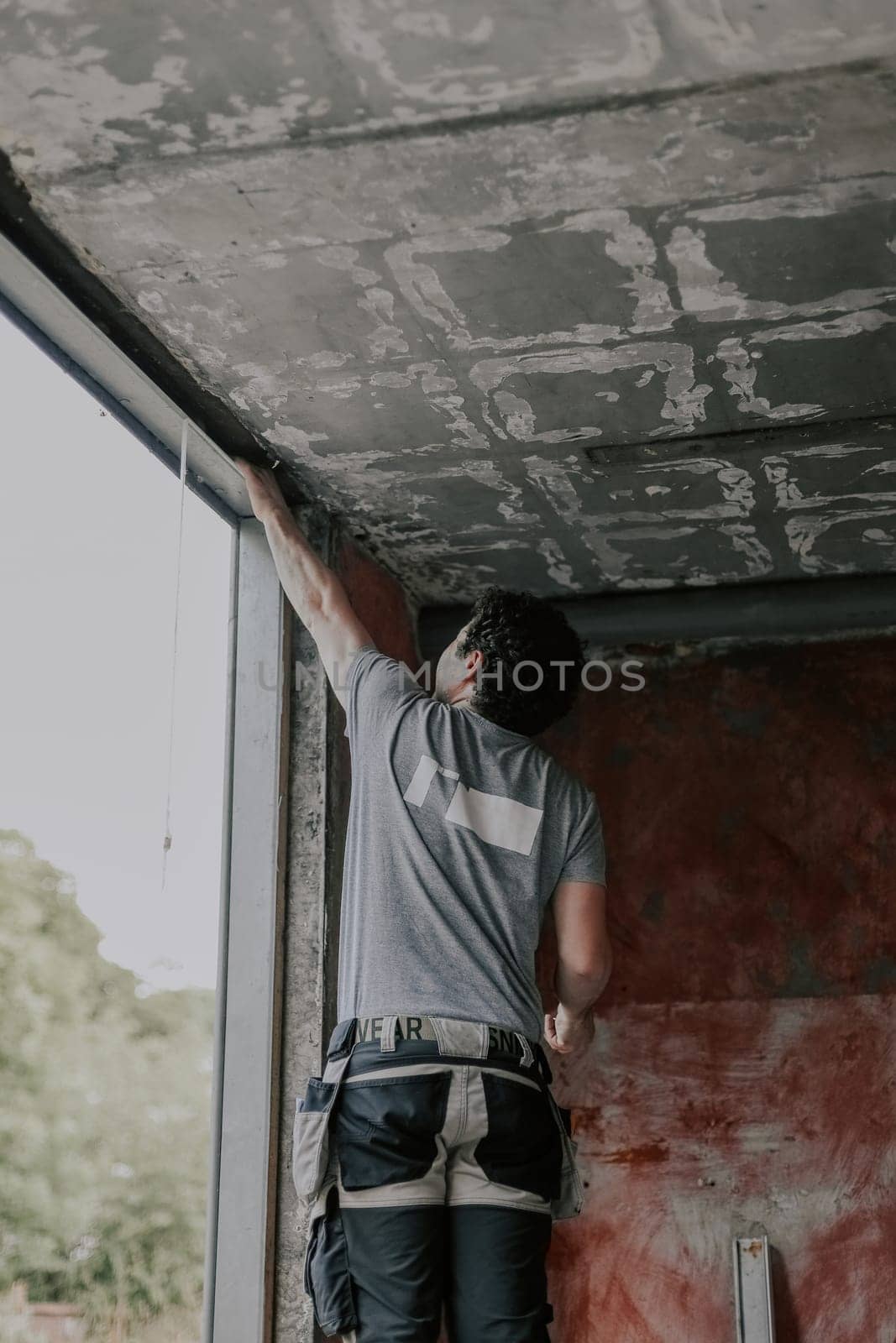 A young Caucasian man in a gray T-shirt with curly brown hair stands on a stepladder and cleans an old window opening with his hand to install a new frame, close-up view from below. The concept of installing windows, construction work, house renovation.