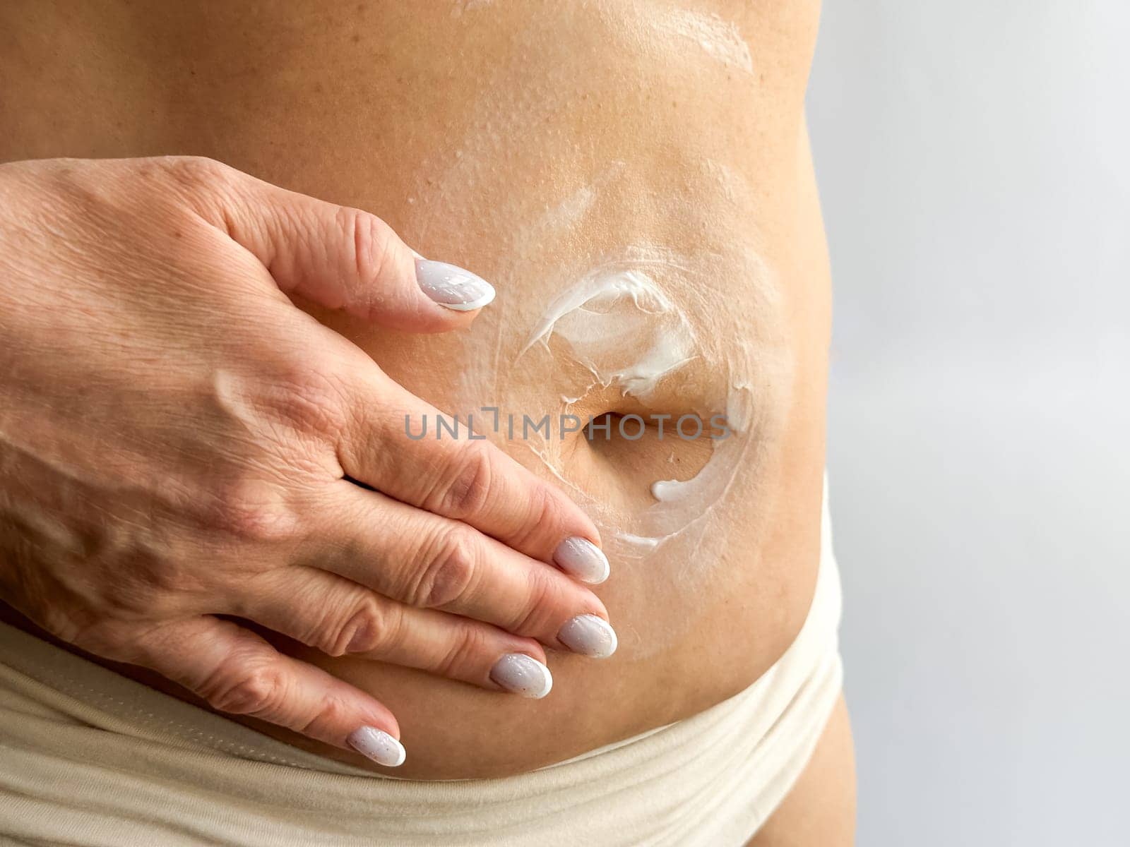 Close up of womans hand with white manicured nails applying cream to her stomach. Highlights skincare, body care, and personal hygiene, focusing on moisturizing and self care routines. High quality
