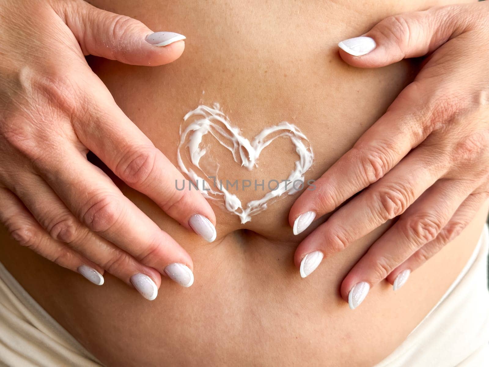 Close up of womans hands middle age with white nails applying cream in the shape of heart on her stomach. Skincare, self care, and personal hygiene with on love and body positivity. by Lunnica