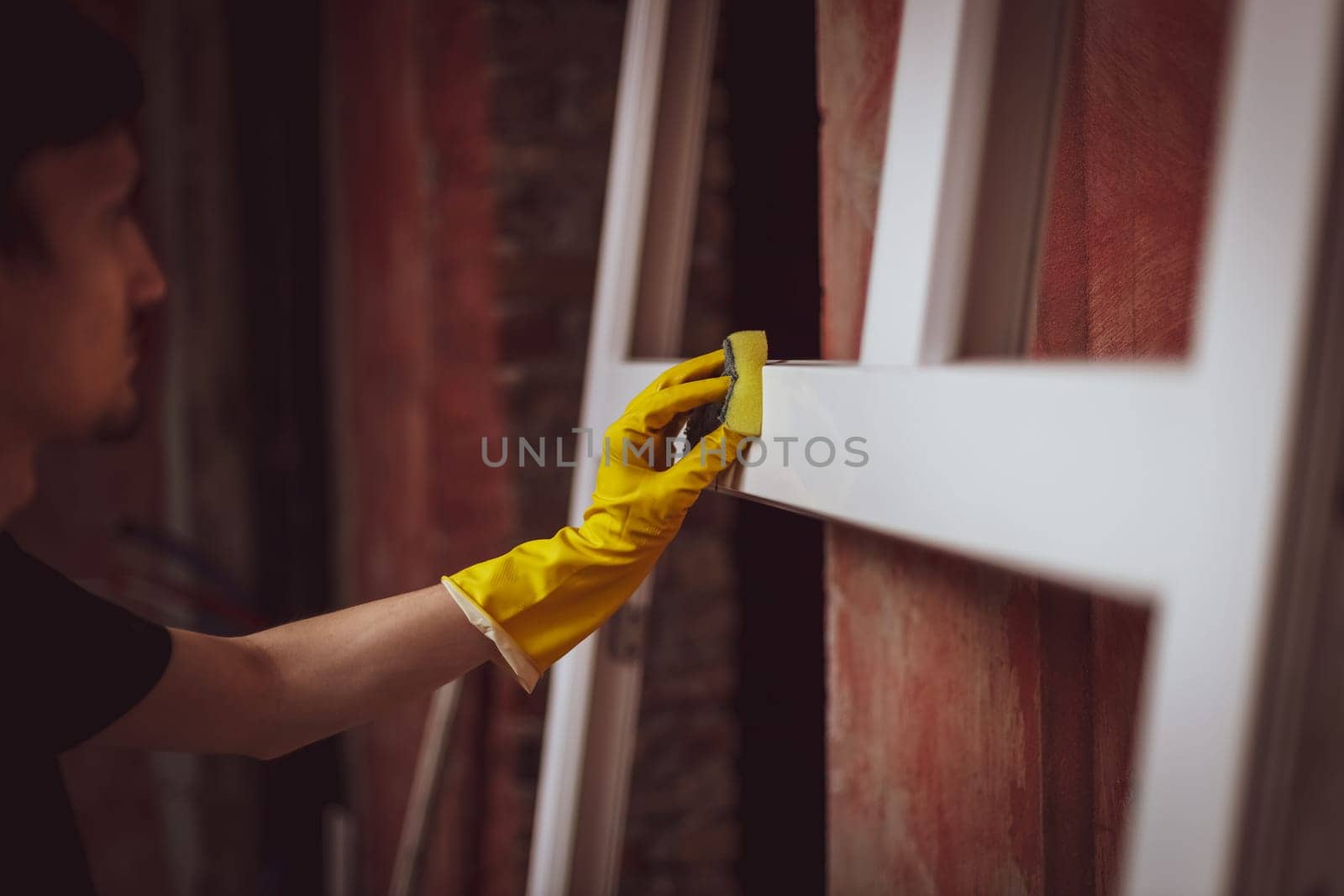 A young caucasian man in yellow gloves cleans a window frame with a sponge and soap, preparing them for installation, close-up side view. The concept of home renovation, installation of windows, construction work.