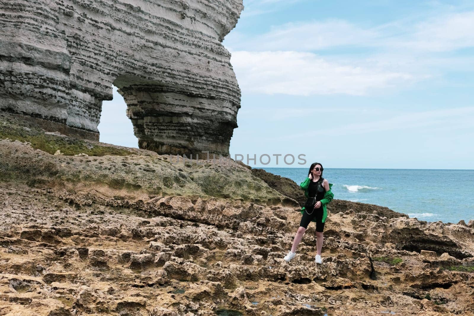 A beautiful caucasian young brunette girl in a green shirt and sunglasses stands posing near the arch on the rocky shore of the north sea in Etretat in France, close-up side view. People and nature concept.