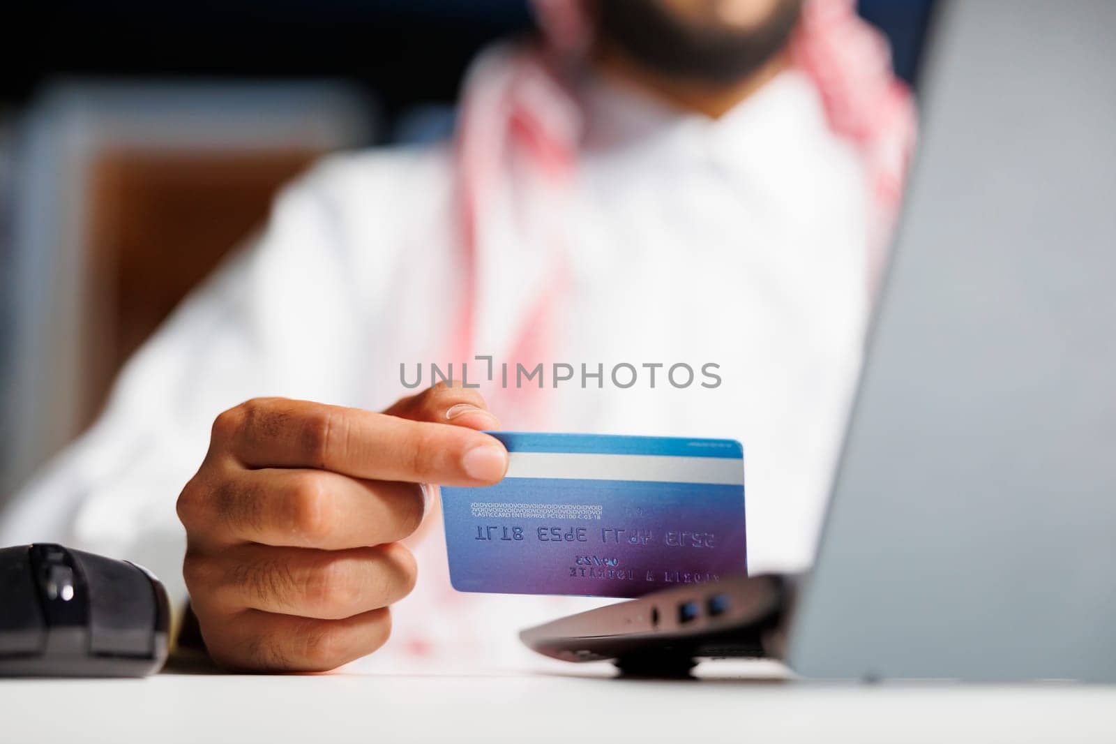 Focused Arab businessman at a modern desk, engrossed in efficient online work. Using wireless technology, he types, researches and shops securely with a credit card.