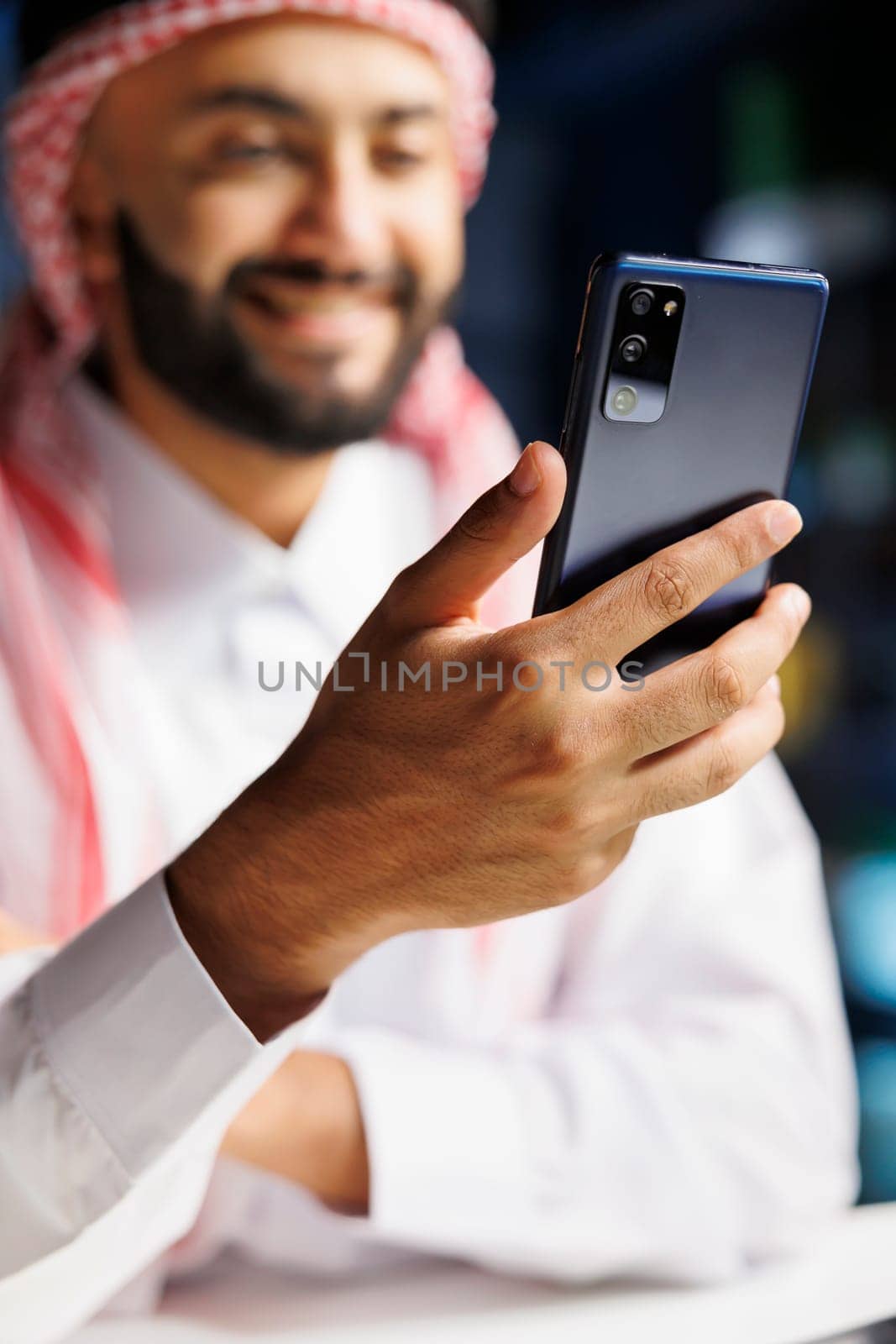 Middle Eastern entrepreneur in traditional clothing sits at a modern desk, browsing the internet on his cell phone. With a smile, an Islamic man uses wireless technology for work. Close-up shot.