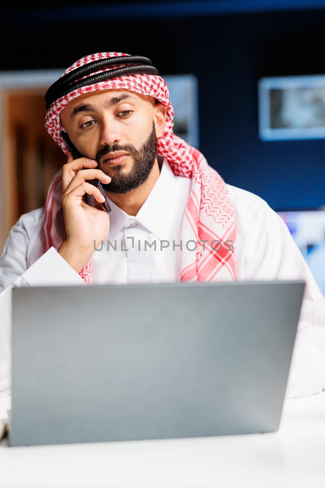 Detailed shot of Arab man utilizing a laptop and mobile device for conversation and study, demonstrating technological aptitude. Multitasking Muslim guy conversing on his mobile and exchanging notes.