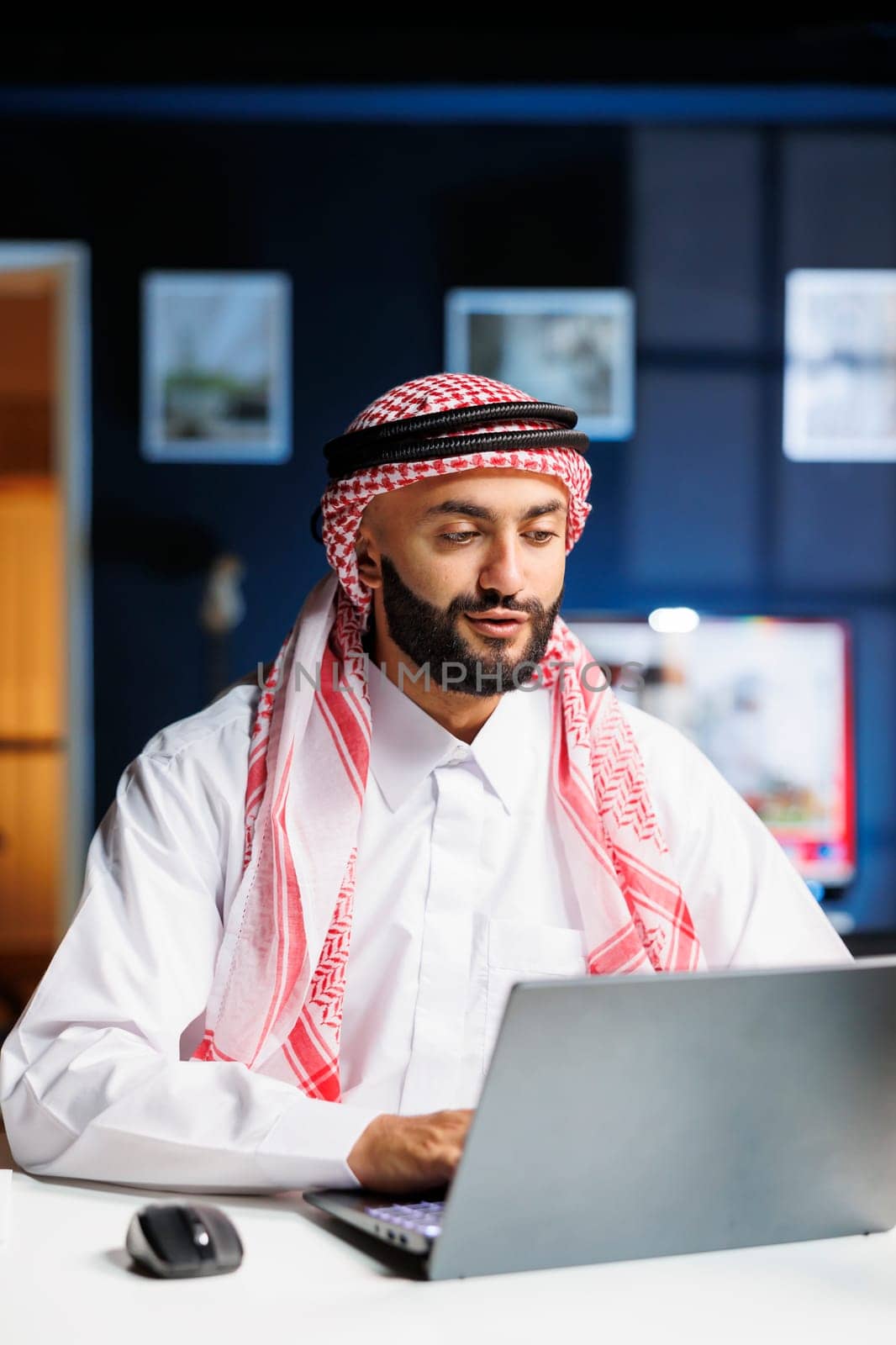 Showcasing professional dedication is Middle Eastern man in traditional clothing utilizing his personal computer for online research. Detailed view of Arab guy diligently working on digital laptop.