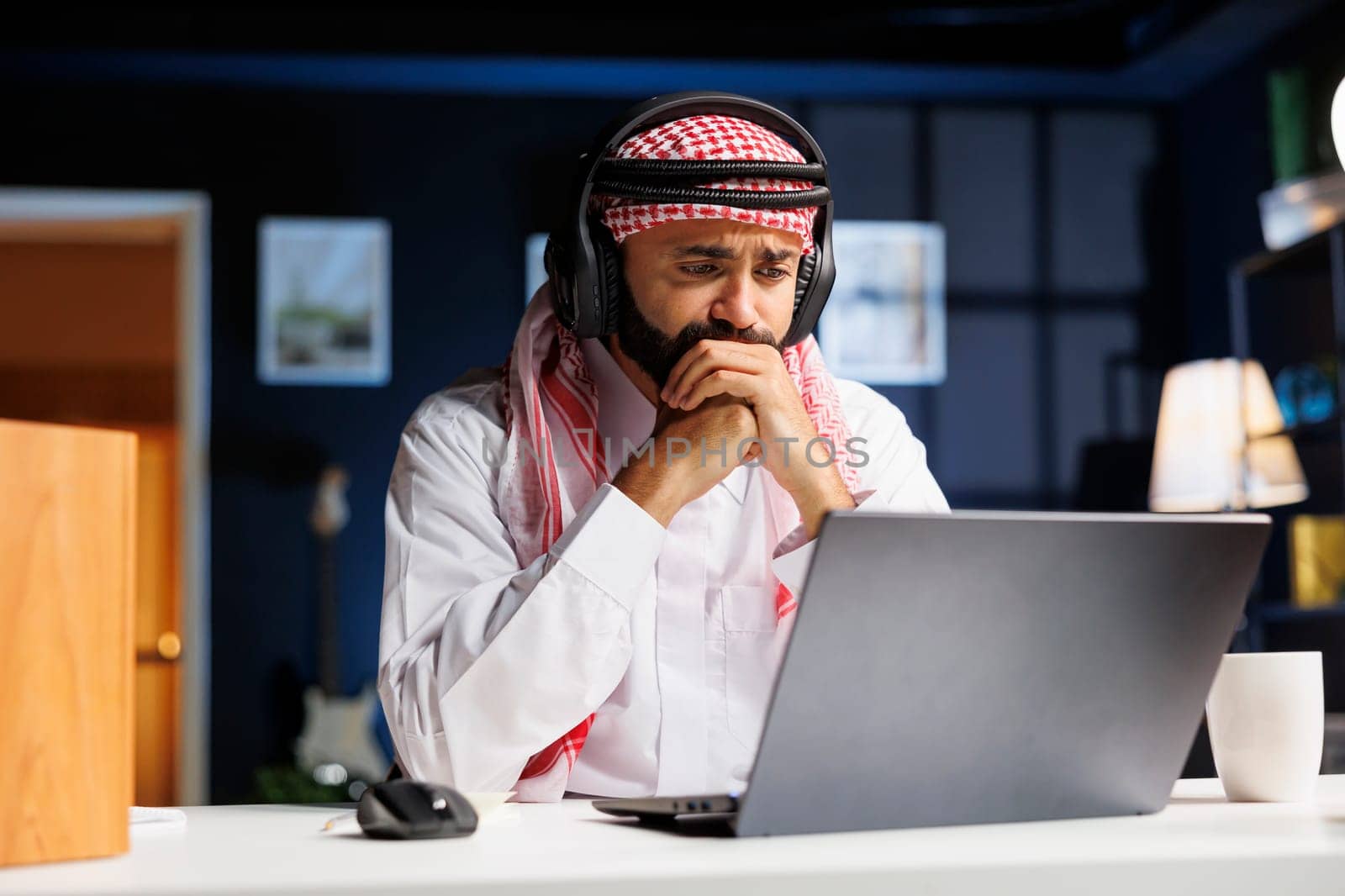 A focused man in traditional Arabic attire works diligently at his desk, utilizing wireless technology. He researches online, communicates, and engages in video conferencing on his personal computer.