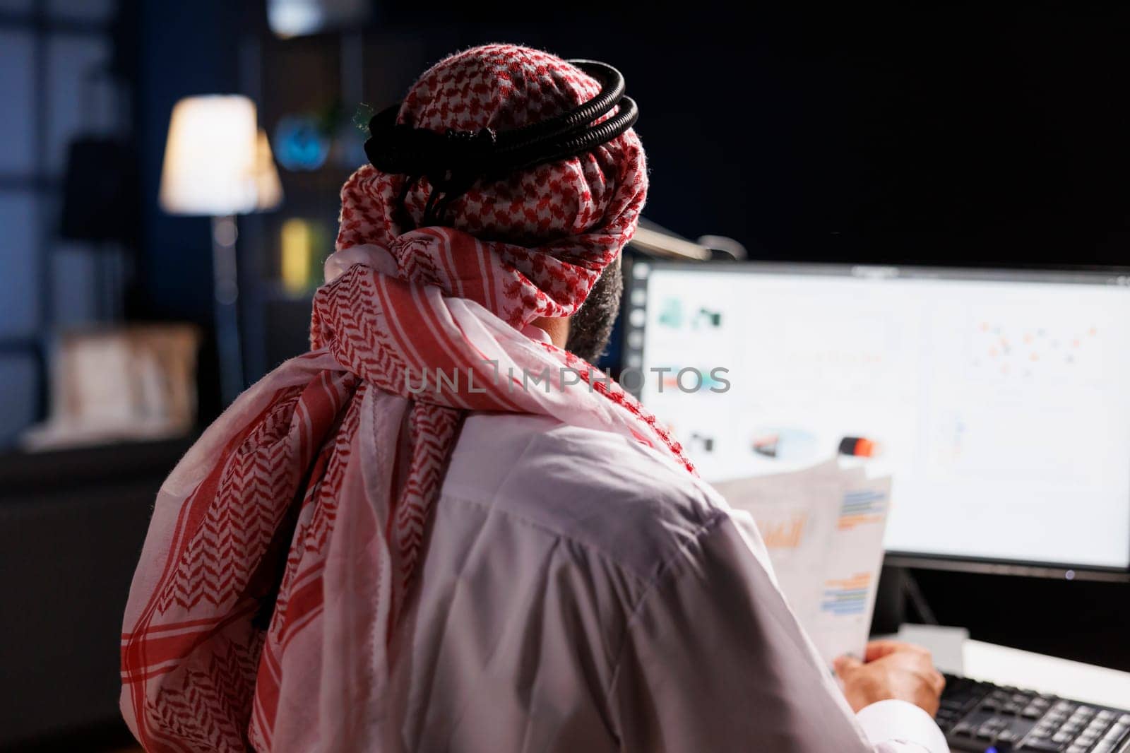 Focused Arab businessman diligently works at his desk, conducting online tasks and examining paperwork. Over-the-shoulder shot of a Muslim guy holding papers and using his modern desktop computer.