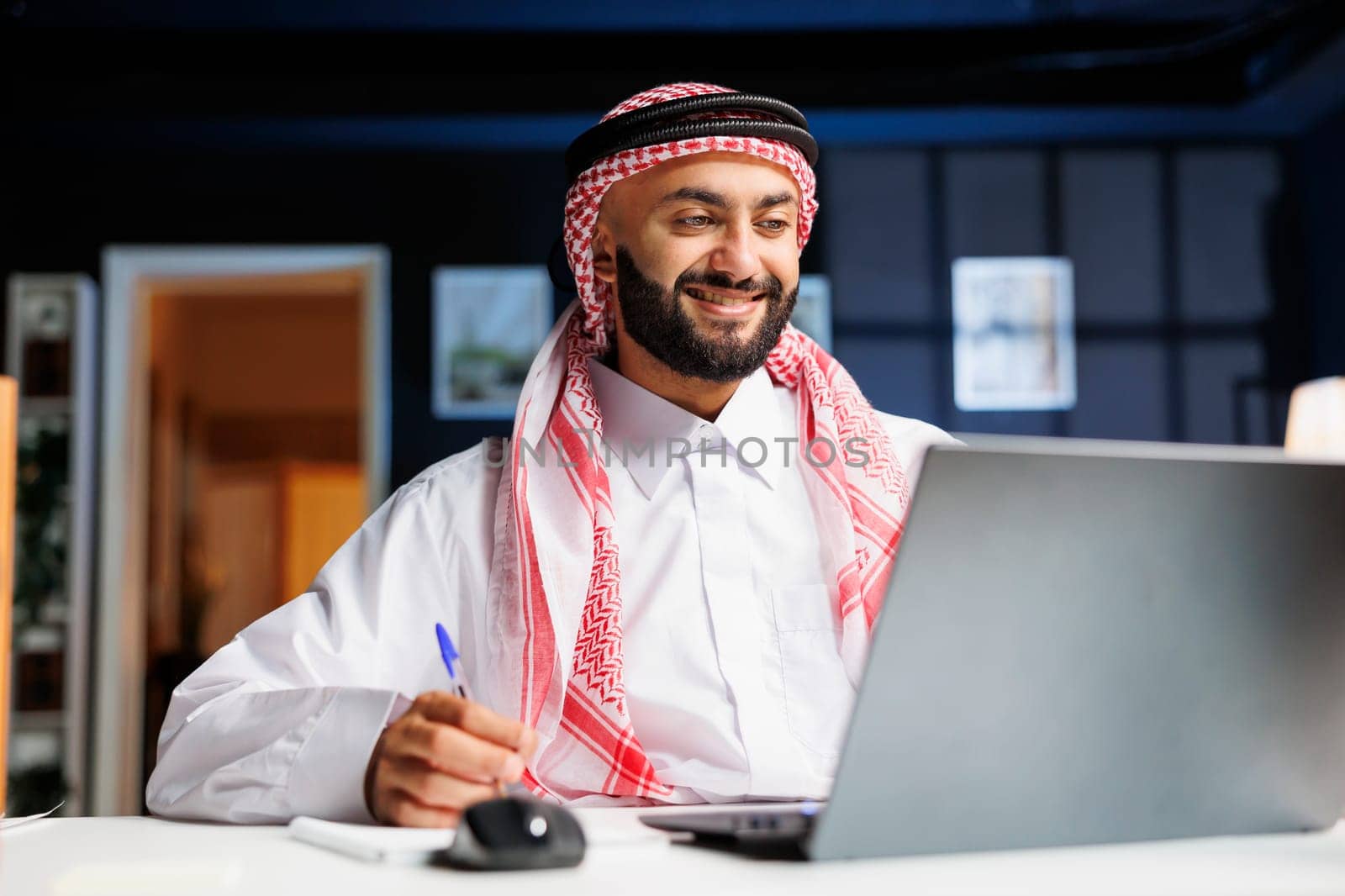 Smiling Arab guy works diligently at office desk, engages in digital tasks and writes on his notepad with laptop nearby. He efficiently conducts online research, embodying dedication and productivity.