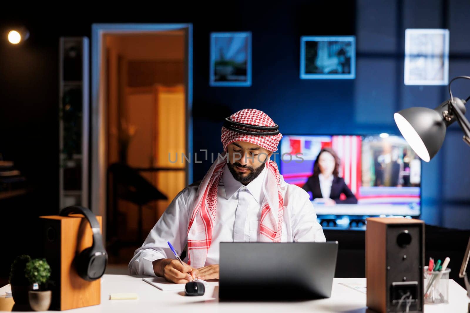 Arab man writing on notebook and using a wireless device for browsing the internet at his modern office desk. Muslim guy doing research by utilizing his digital personal computer.