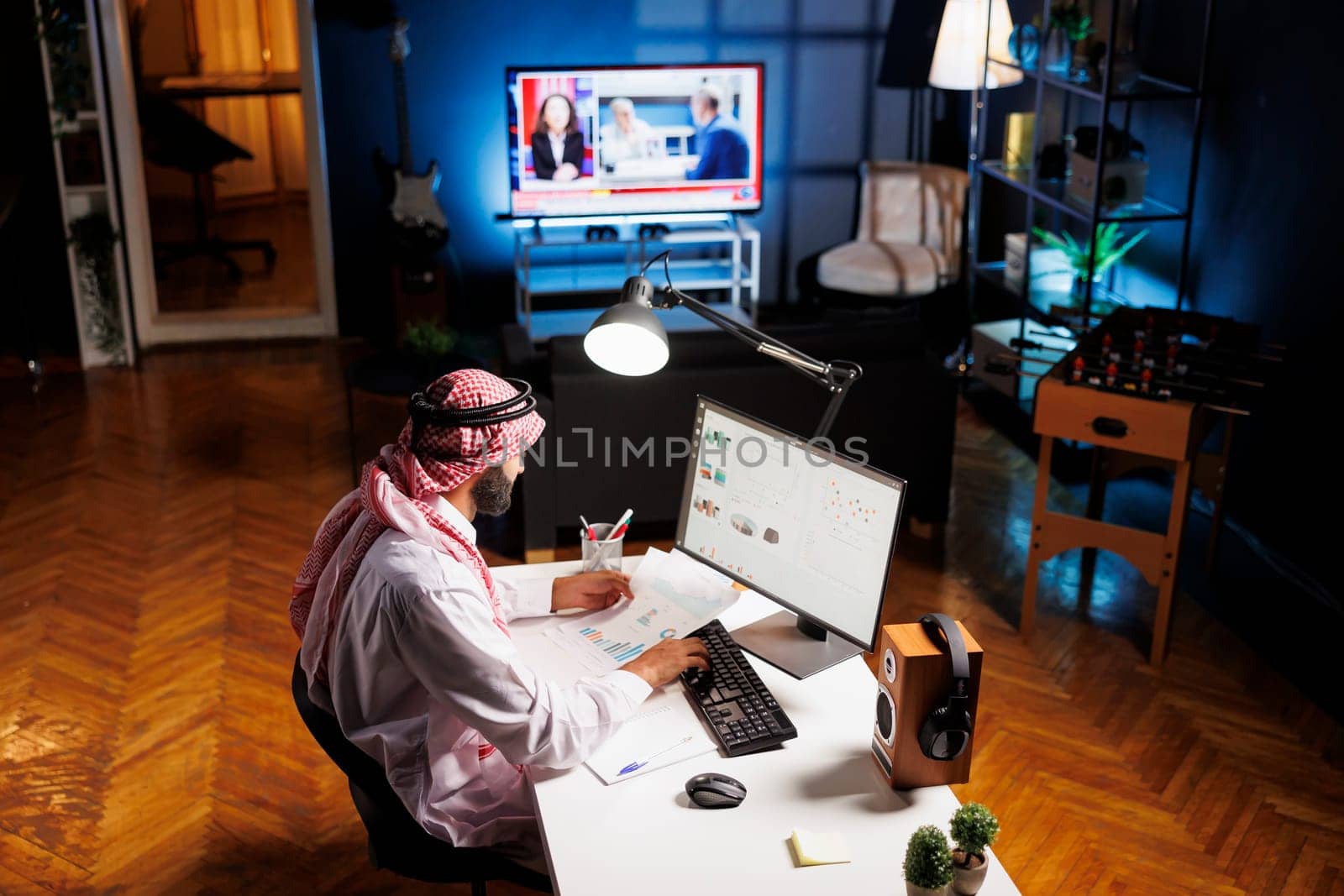 Muslim guy with paperwork diligently conducting online tasks efficiently. Young man in Islamic clothing manages his work with dedication and meticulousness by using a desktop PC. Overhead shot.