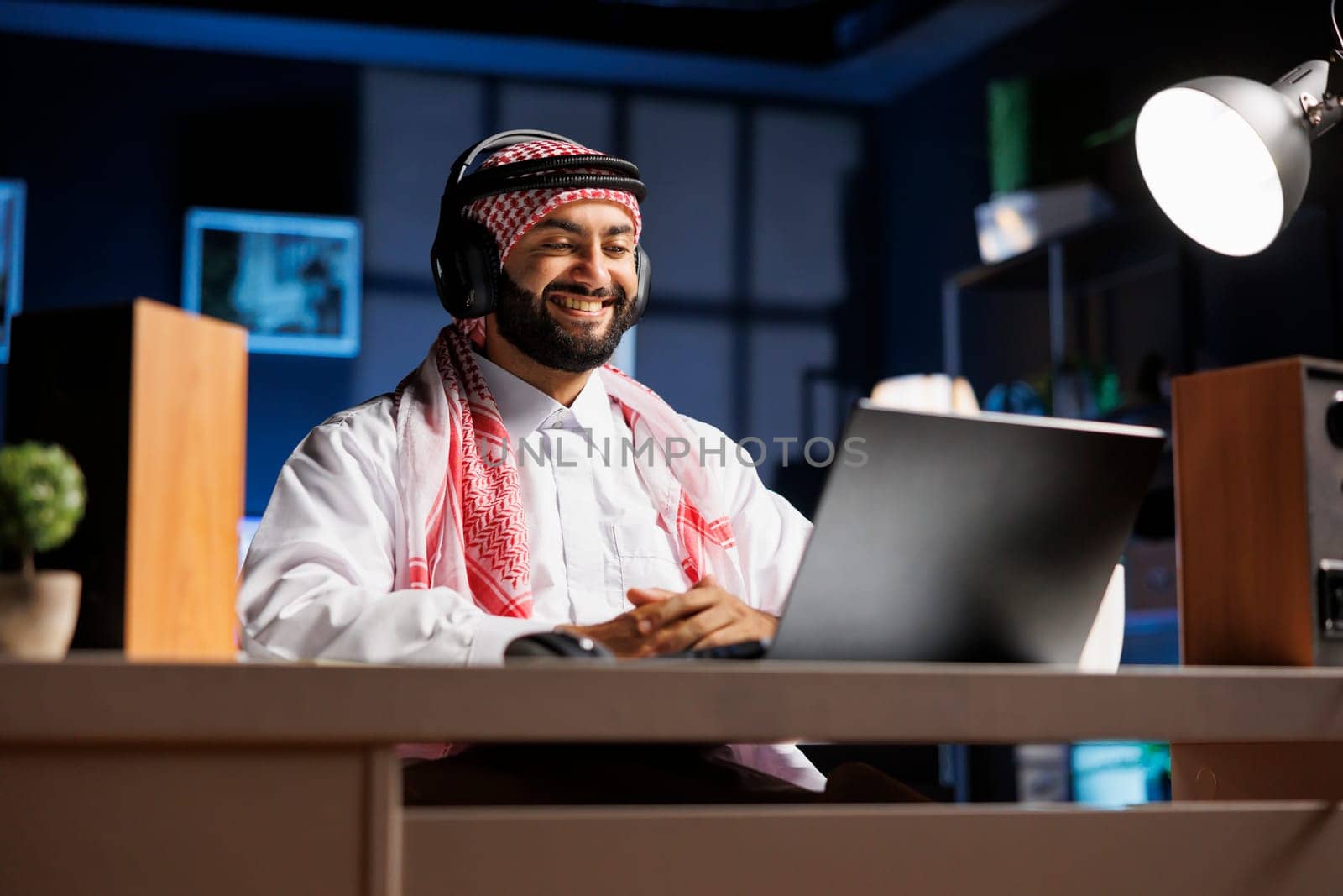 Handsome man in traditional Islamic attire, sitting at desk with laptop, smiles as he does research online. Arab guy with wireless headphones engrossed in video conferencing and email communication.