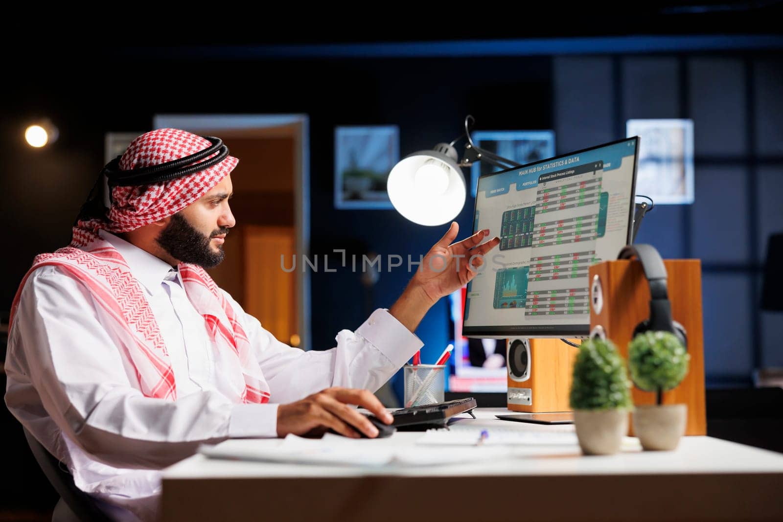 Handsome male professional focusing on work in a well-organized office space, surrounded by technology and paperwork. Muslim man closely examining the data research on pc monitor.