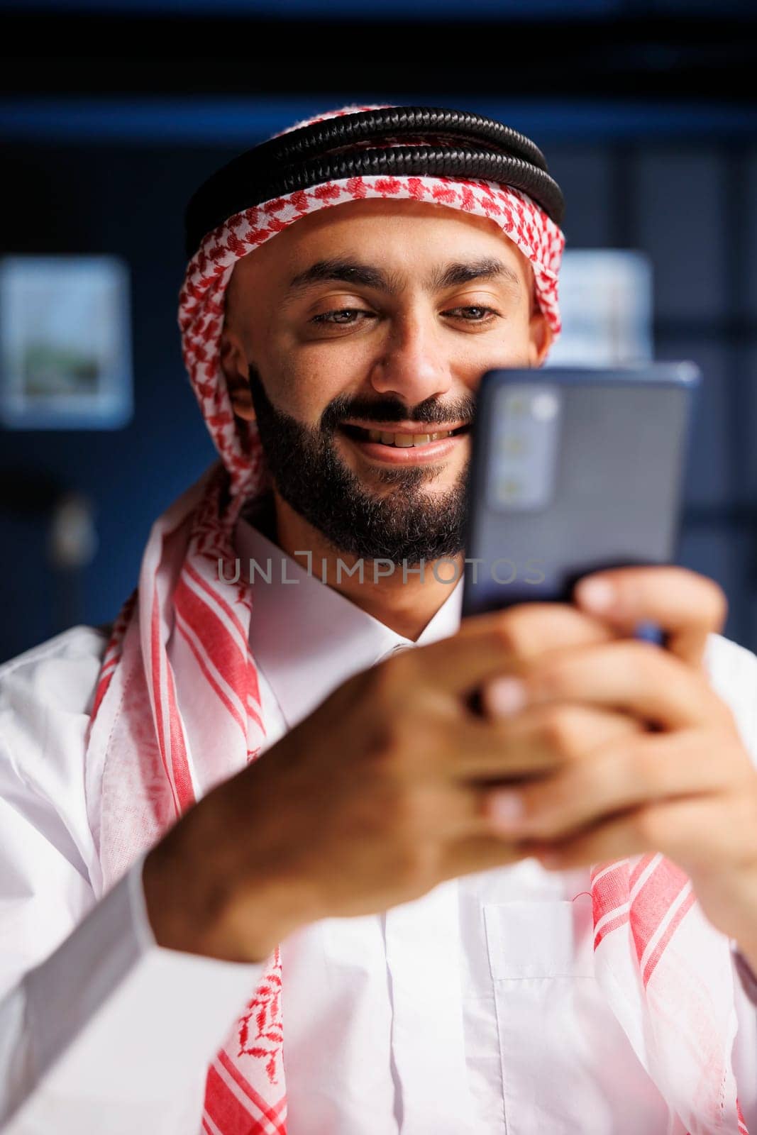 Detailed portrait shot of a smiling Muslim man using a smartphone for digital communication. Close-up view of an Arab entrepreneur typing and researching on his mobile device.