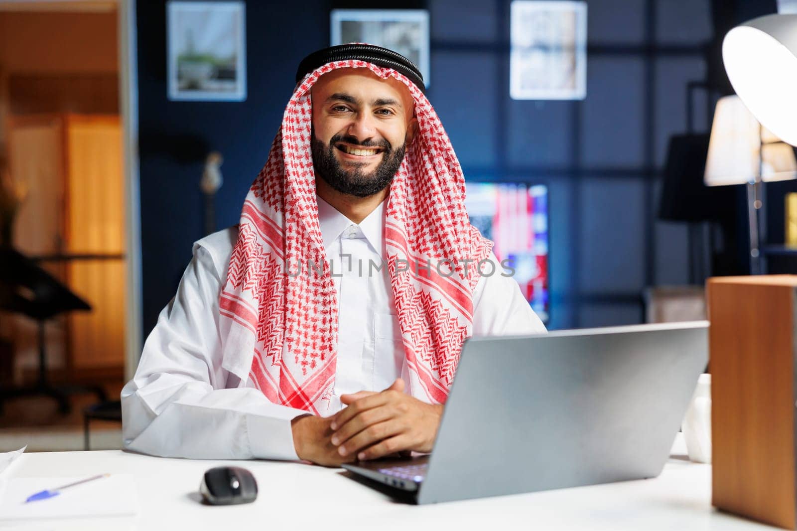 Male person dressed in traditional Arabic clothing is seated with his laptop. Confident and tech-savvy, the Arab man smiles at the camera, embodying the entrepreneurial spirit of the digital age.