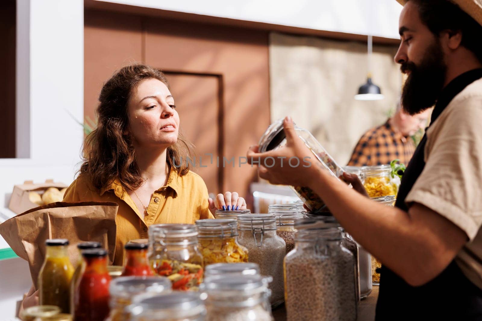 Woman shopping in zero waste grocery store, looking at bulk products in reusable packaging while being assisted by smiling seller. Customer in local neighborhood shop receiving free food sample