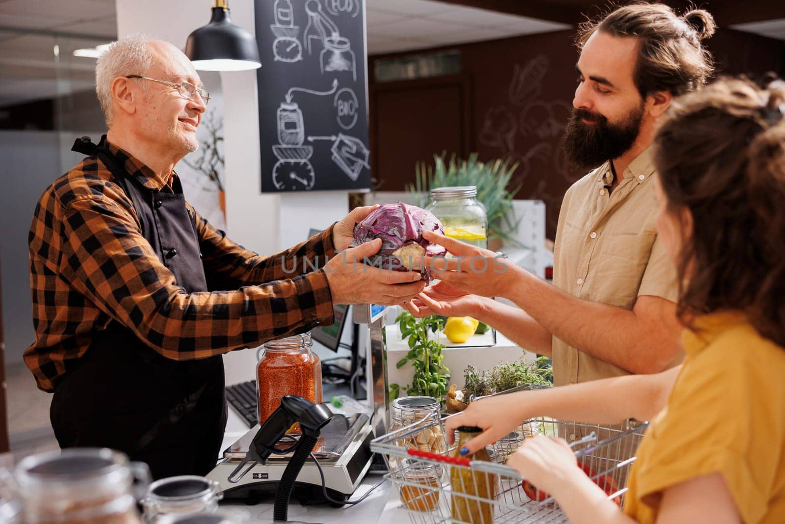 Older man at checkout counter in zero waste shop selling healthy vegetables in jar to vegan customers with green living lifestyle. Hipster couple buying groceries in local neighborhood store
