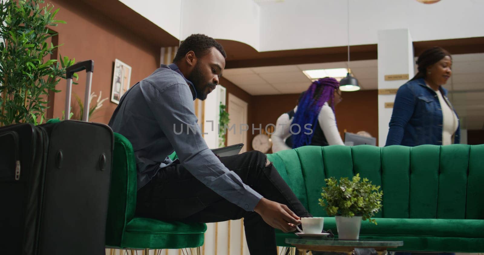 Hotel guest browsing tablet and enjoying coffee while he waits on room reservation check in, relaxing in lounge area at holiday resort. Young traveller receiving luxurious concierge services.