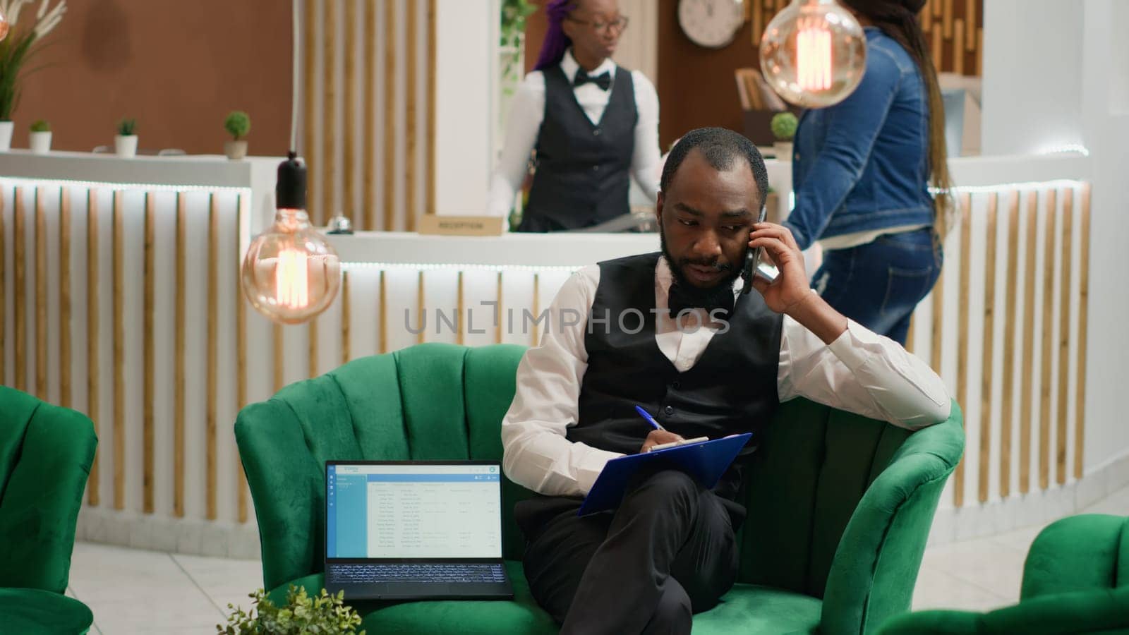 Bellboy talking on phone and taking notes, sitting in lobby at five star hotel. Concierge calling guests to ensure easy check in with all personal information, assisting visitors in arranging room.