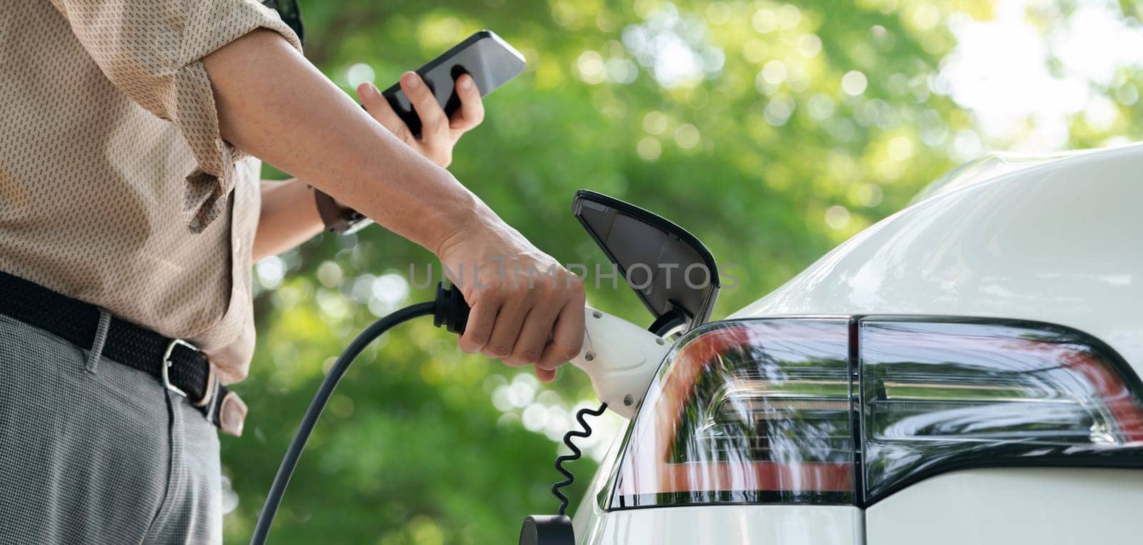 Young man use smartphone to pay for electricity at public EV car charging station green city park. Modern environmental and sustainable urban lifestyle with EV vehicle. Panorama Expedient