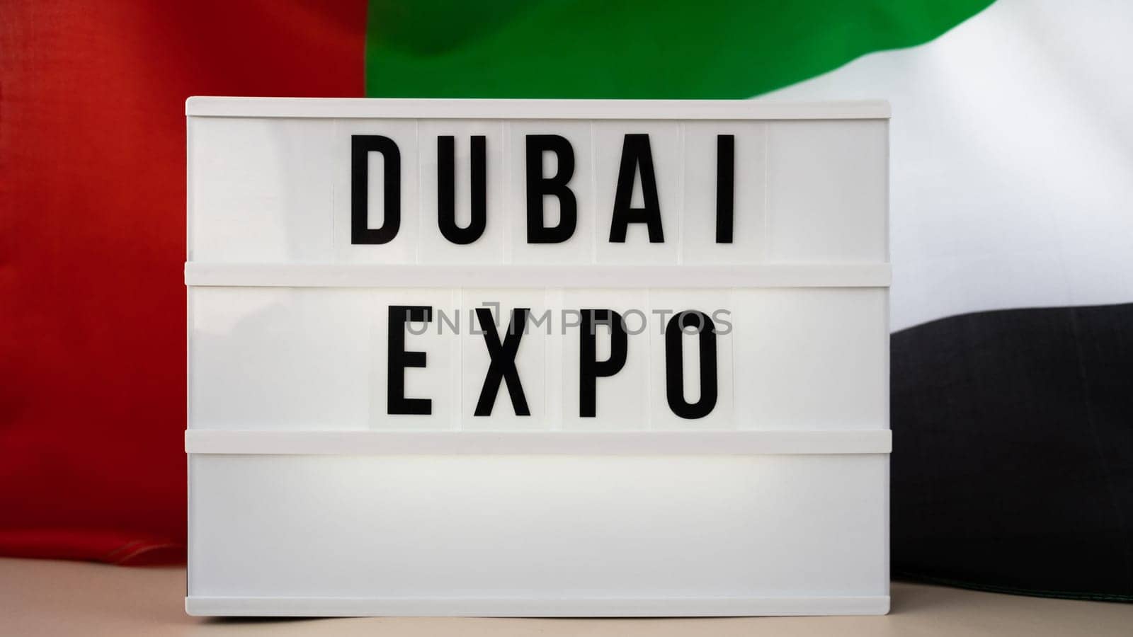 Lightbox with text DUBAI EXPO on background of waving UAE flag made from silk. United Arab Emirates flag with concept of tourism and traveling. Inviting greeting card, advertisement. Dubai welcoming card