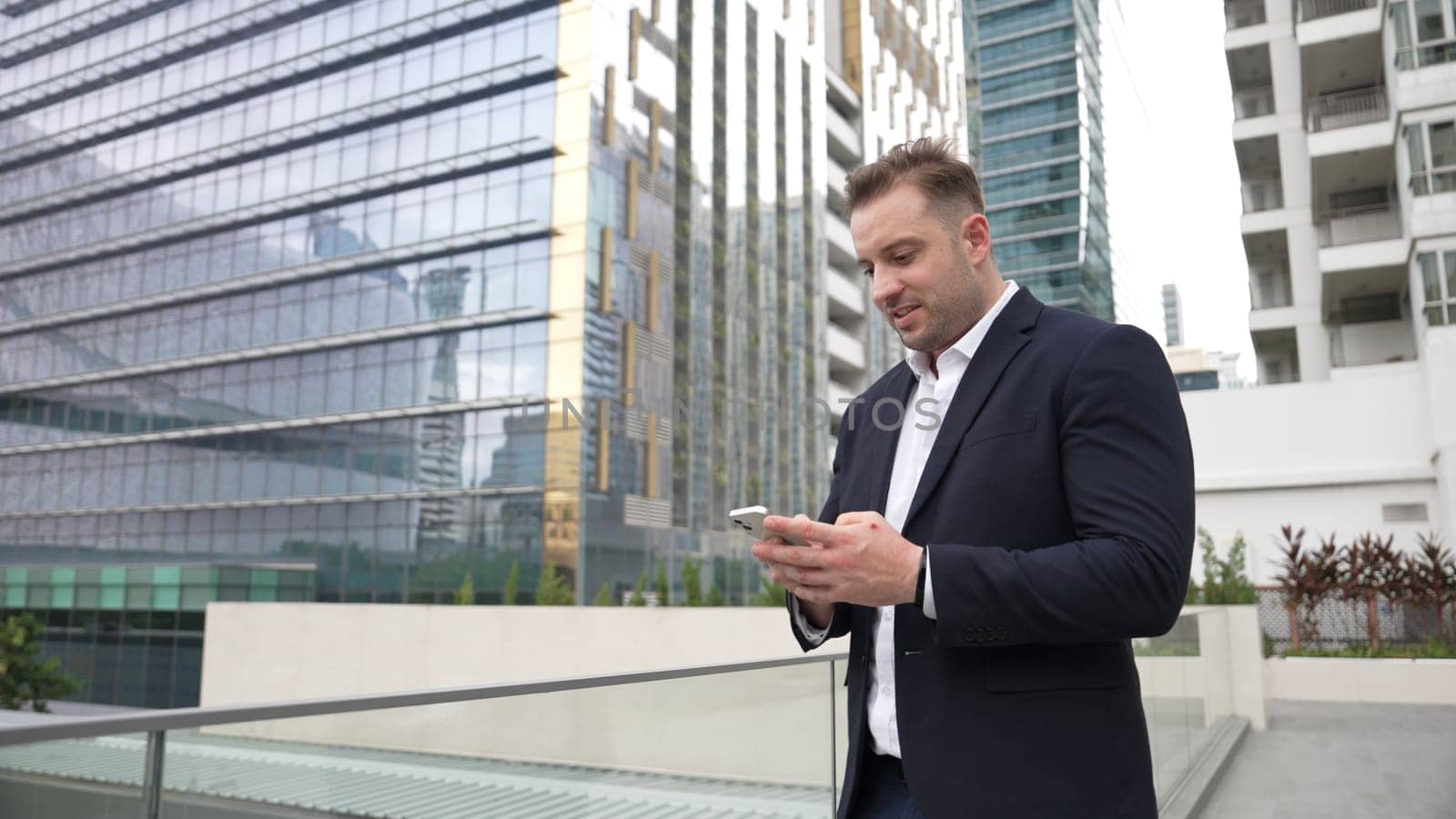 Happy businessman using phone to chatting with team while standing at urban city. Front view of project manager working on telephone with skyscraper view while communicate with marketing team. Urbane.