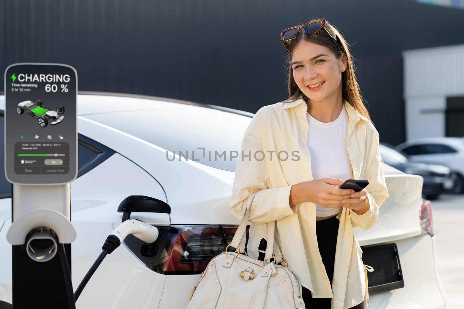 Young woman holding shopping bag and use smartphone to pay for electricity for recharging EV car battery from charging station at city mall parking lot. Modern woman go shopping by eco car. Expedient