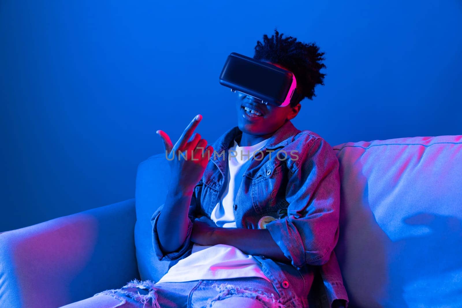 Young African American looking VR enjoy watching in scary funny fantastic 3D cartoon movie technology blue neon light virtual reality metaverse new world at living room sitting lean sofa. Contrivance.