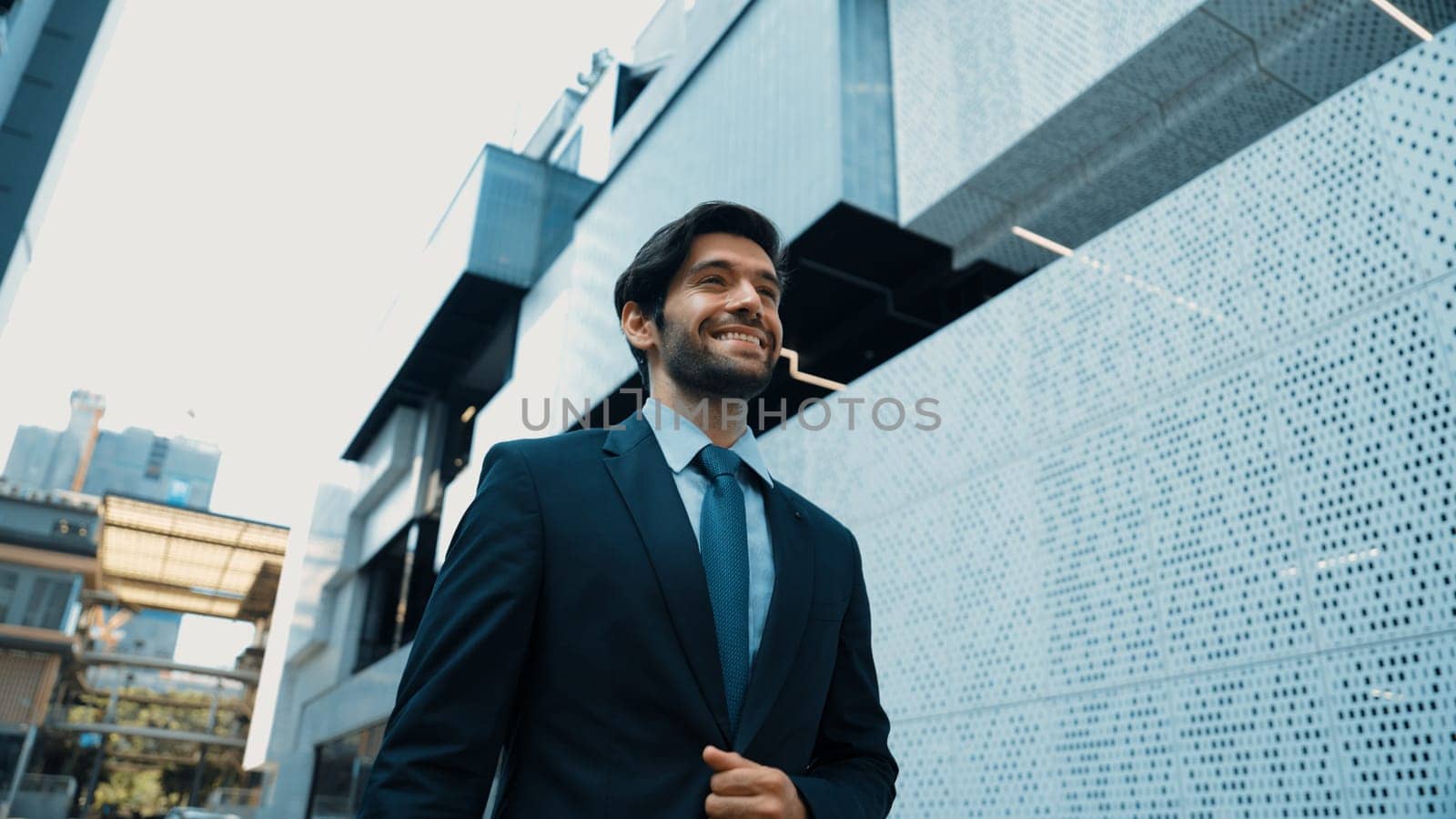 Smart business man wearing suit and smiling while walking at street near white modern architectural building. Attractive handsome manager looking skyscraper in urban city. Copy space. Exultant.