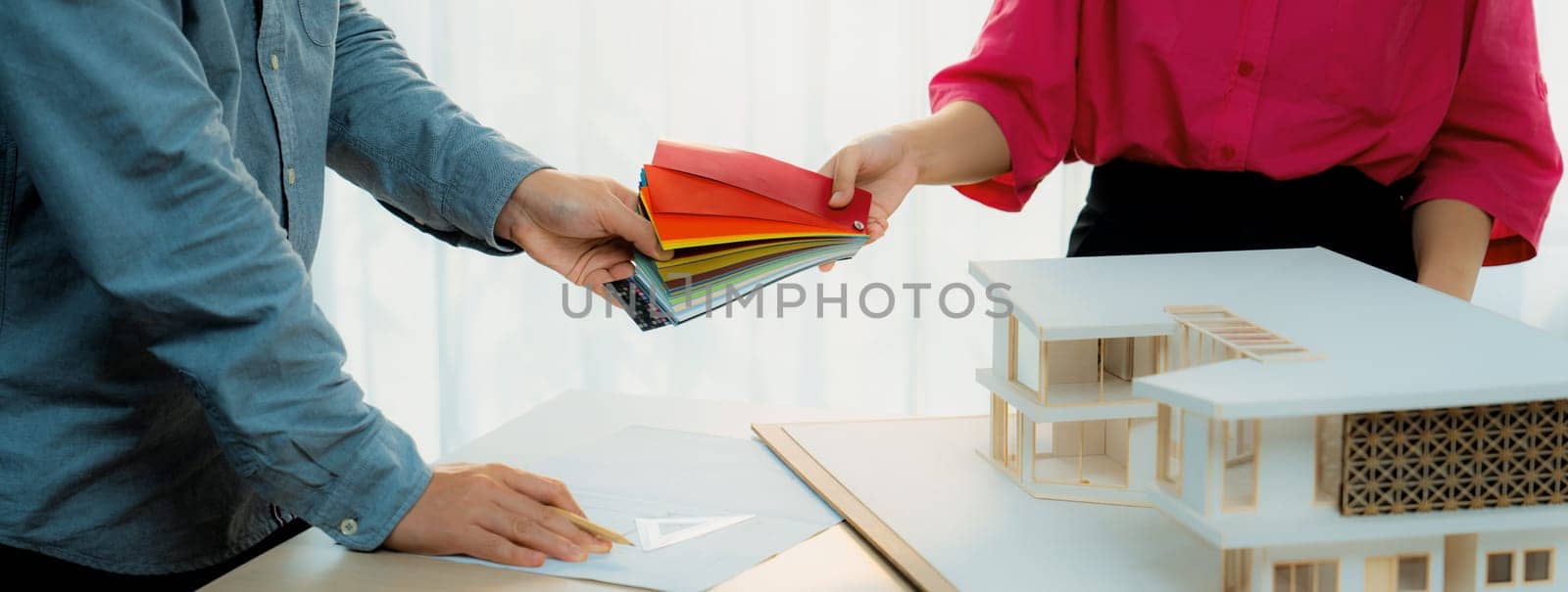 Female architect interior designer send color swatches to project manager for choosing an appropriate color with house model and architectural equipment placed on meeting table. Variegated.