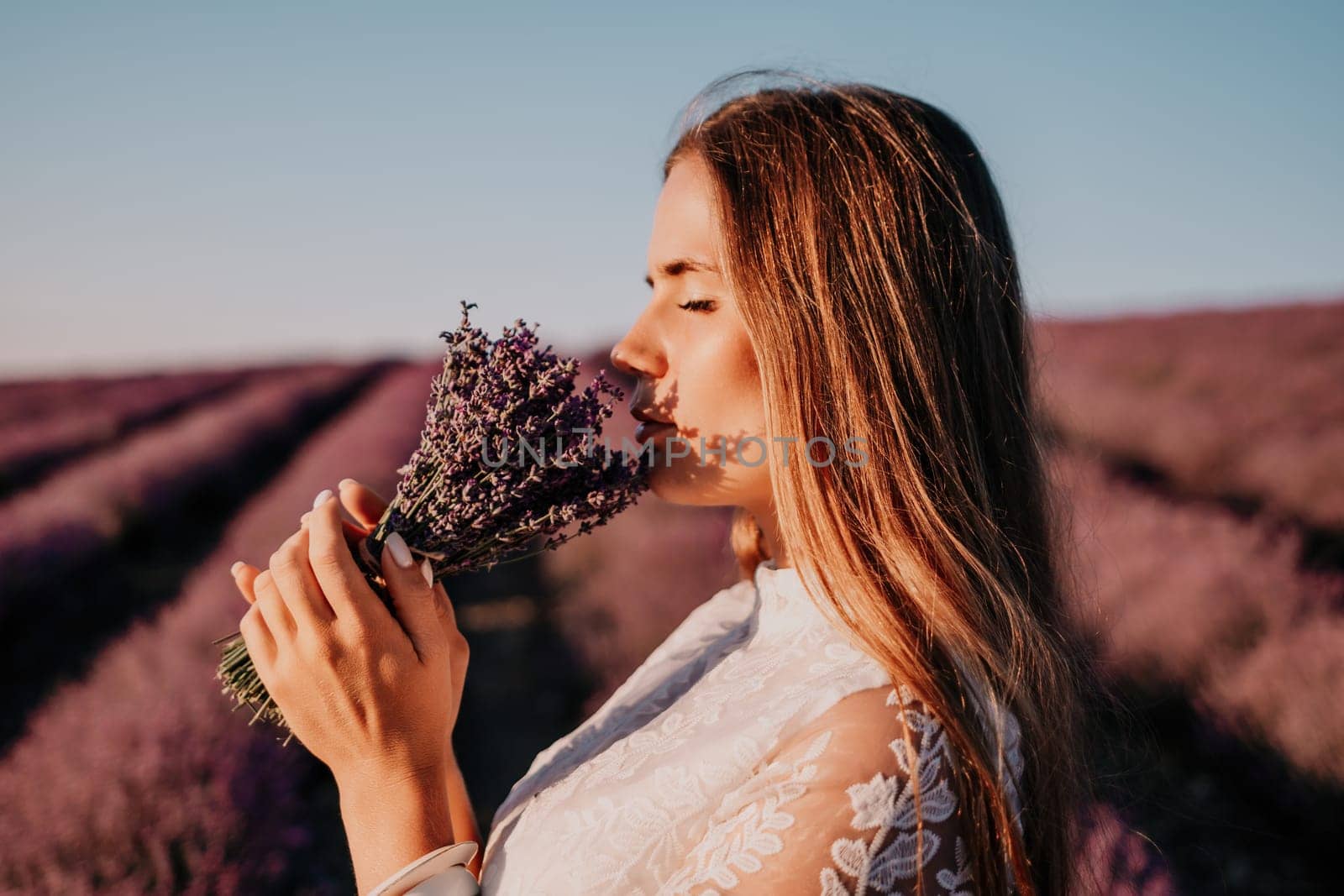 Close up portrait of young beautiful woman in a white dress and a hat is walking in the lavender field and smelling lavender bouquet.