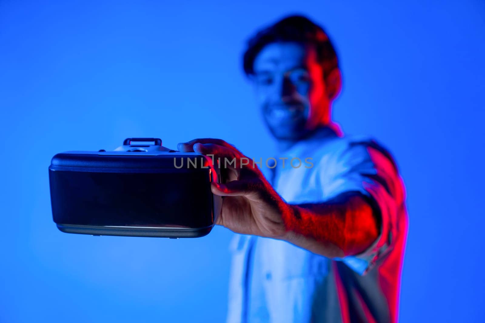 Smiling caucasian man holding and show VR headset with blurring background while standing with neon light. Smart teenager present digital goggles to enter virtual reality game or metaverse. Deviation.