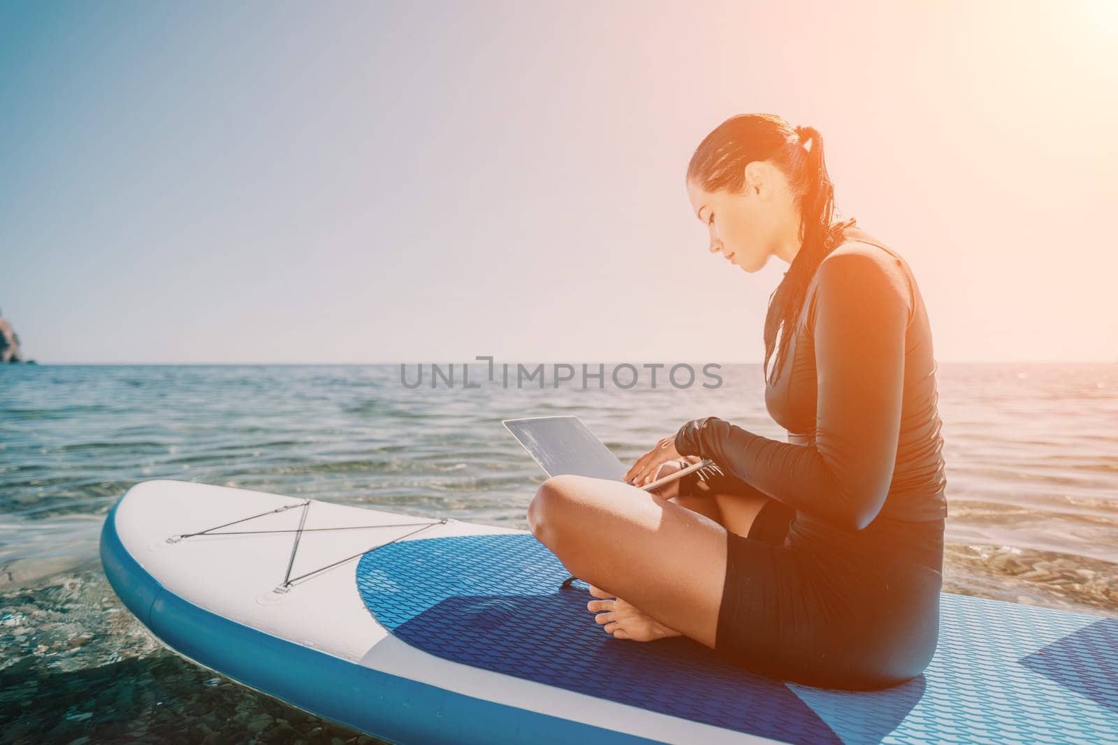 Digital nomad, woman in the hat, a business woman with a laptop sits on the rocks by the sea during sunset, makes a business transaction online from a distance. Freelance, remote work on vacation.