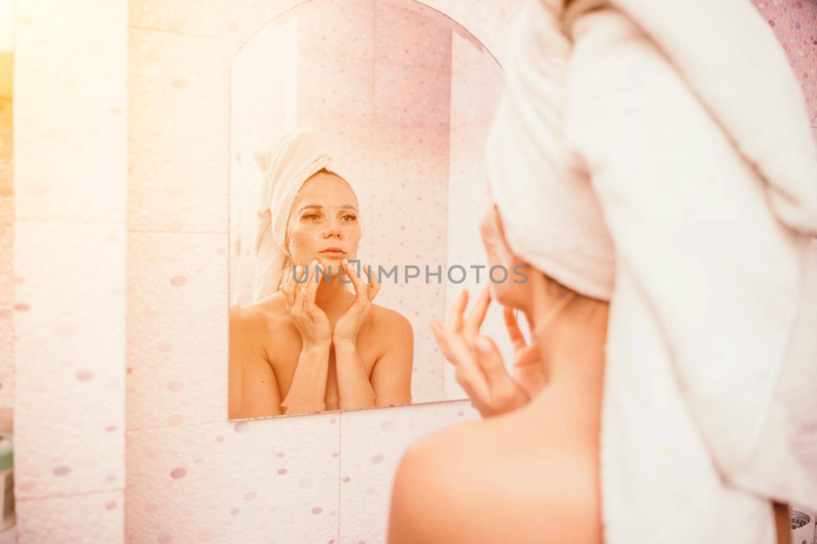 Young beautiful woman using a moisturizing facial mask after taking a bath. Pretty attractive girl in a towel on her head stands in front of a mirror in a home bathroom. Daily hygiene and skin care.