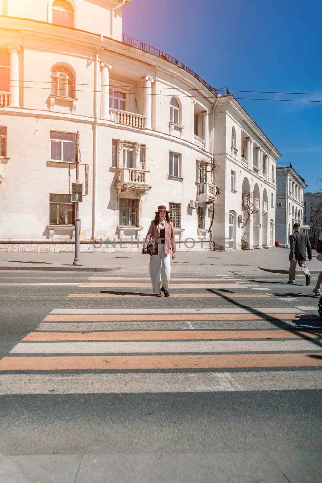 Woman city road crossing. Stylish woman in a hat crosses the road at a pedestrian crossing in the city. Dressed in white trousers and a jacket with a bag in her hands. by Matiunina