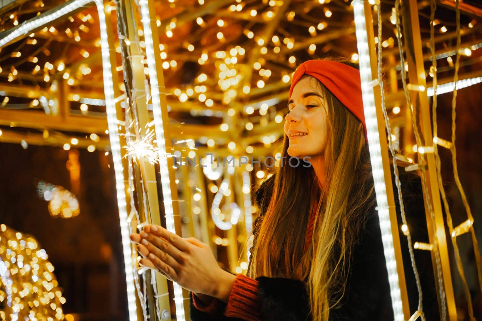 Woman holding sparkler night while celebrating Christmas outside. Dressed in a fur coat and a red headband. Blurred christmas decorations in the background. Selective focus.