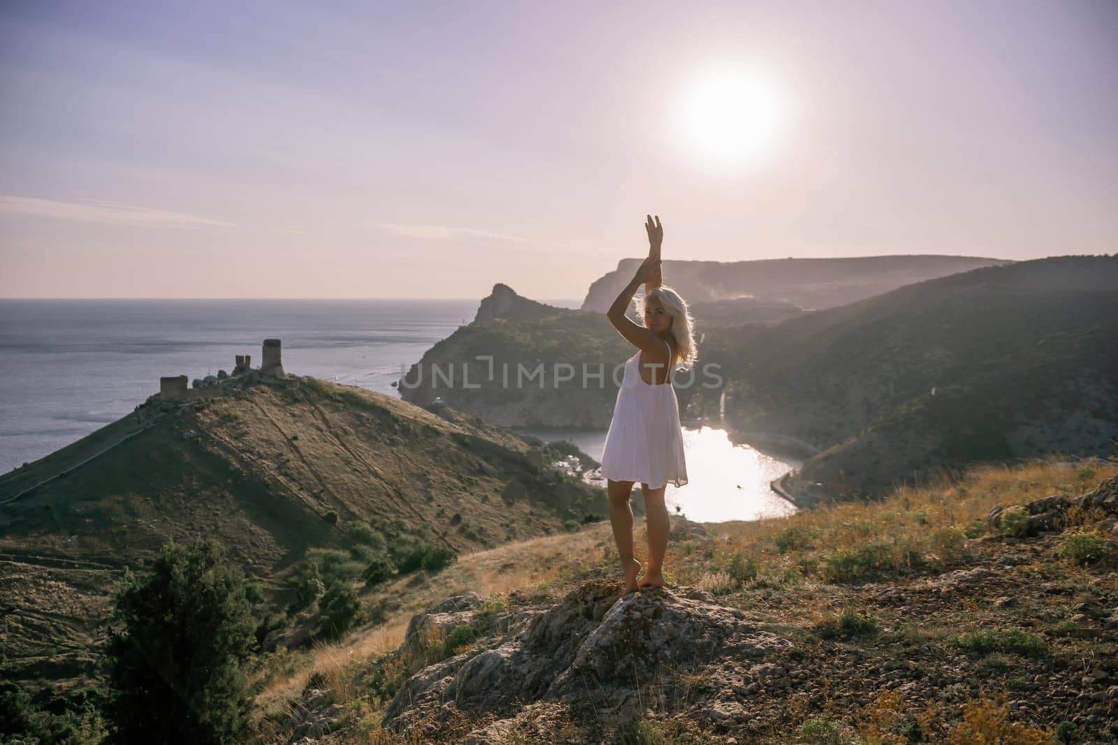 A woman stands on a hill overlooking a body of water. She is wearing a white dress and she is enjoying the view