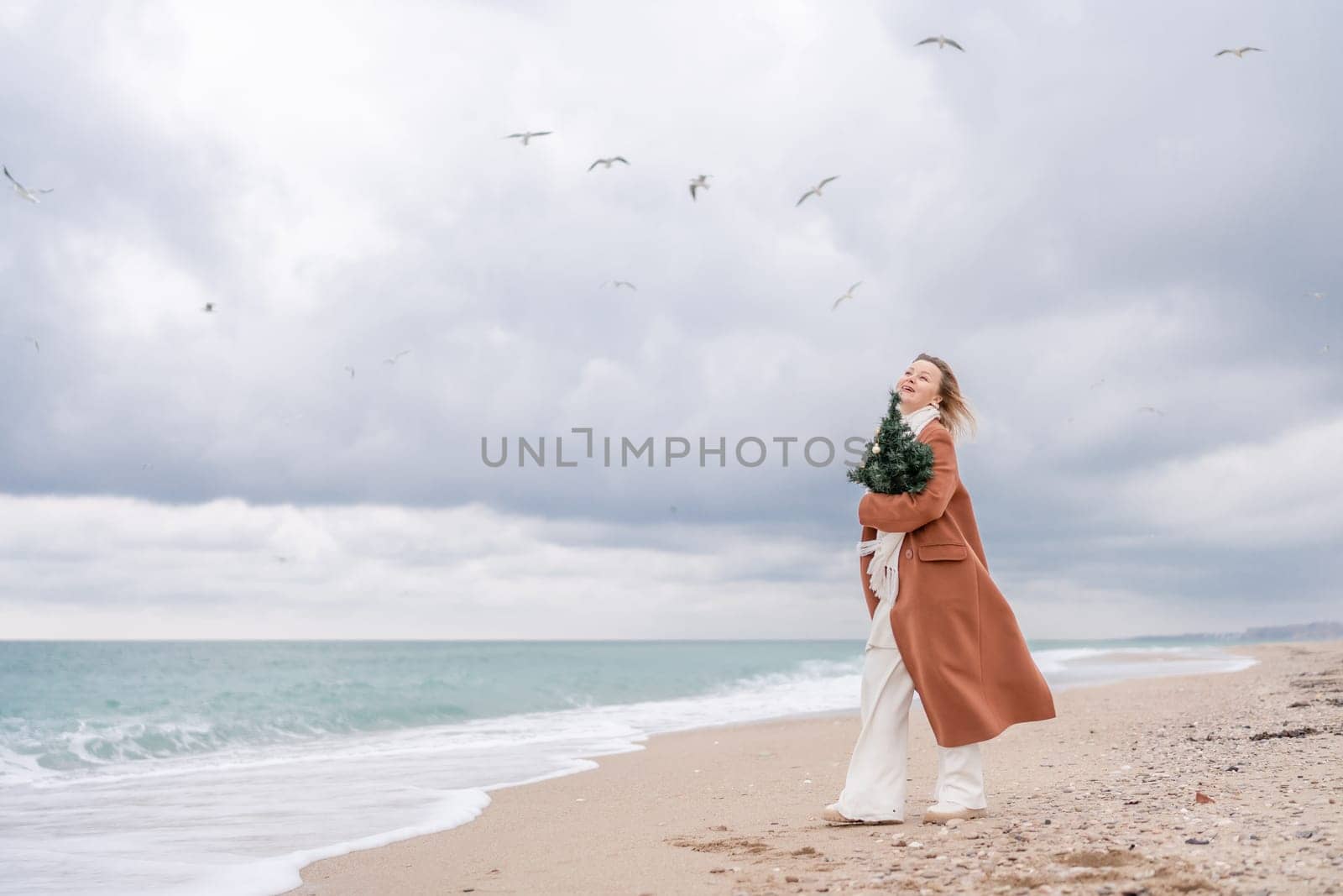 Blond woman Christmas sea. Christmas portrait of a happy woman walking along the beach and holding a Christmas tree on her shoulder. She is wearing a brown coat and a white suit