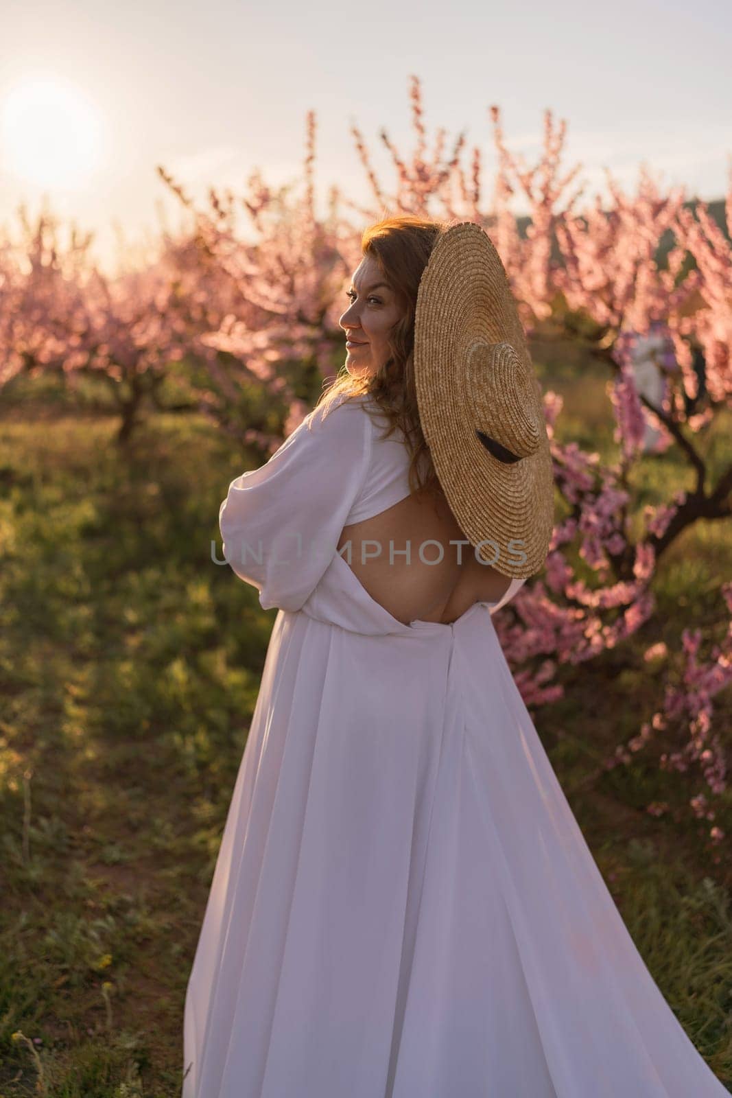 Woman blooming peach orchard. Against the backdrop of a picturesque peach orchard, a woman in a long white dress and hat enjoys a peaceful walk in the park, surrounded by the beauty of nature