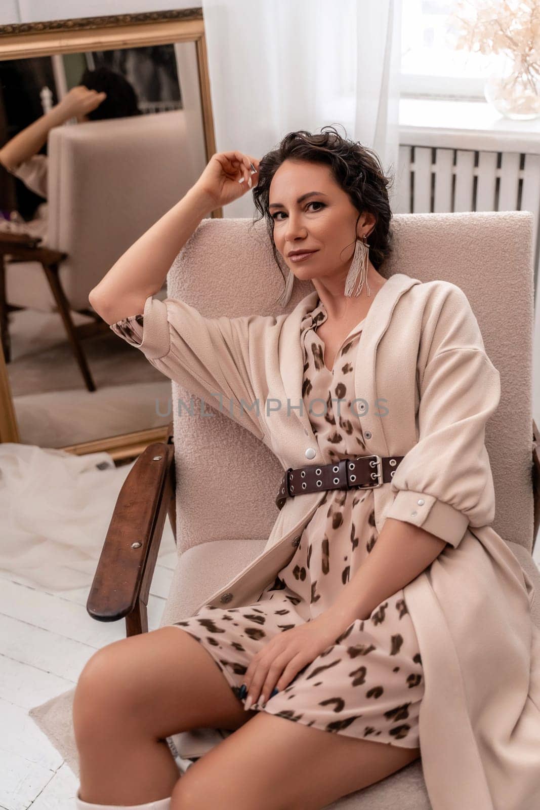 A woman is sitting in a chair wearing a tan jacket and a leopard print dress. She is wearing a belt and earrings