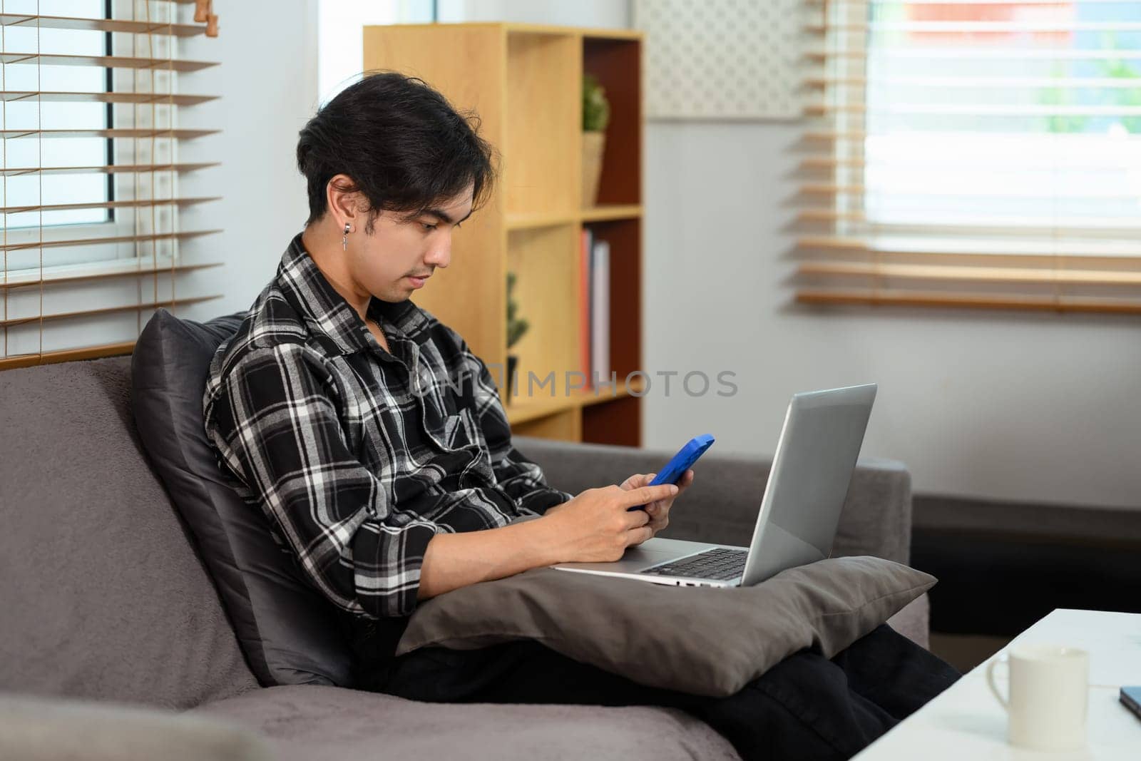 Focused young man using laptop and smartphone on couch in living room.