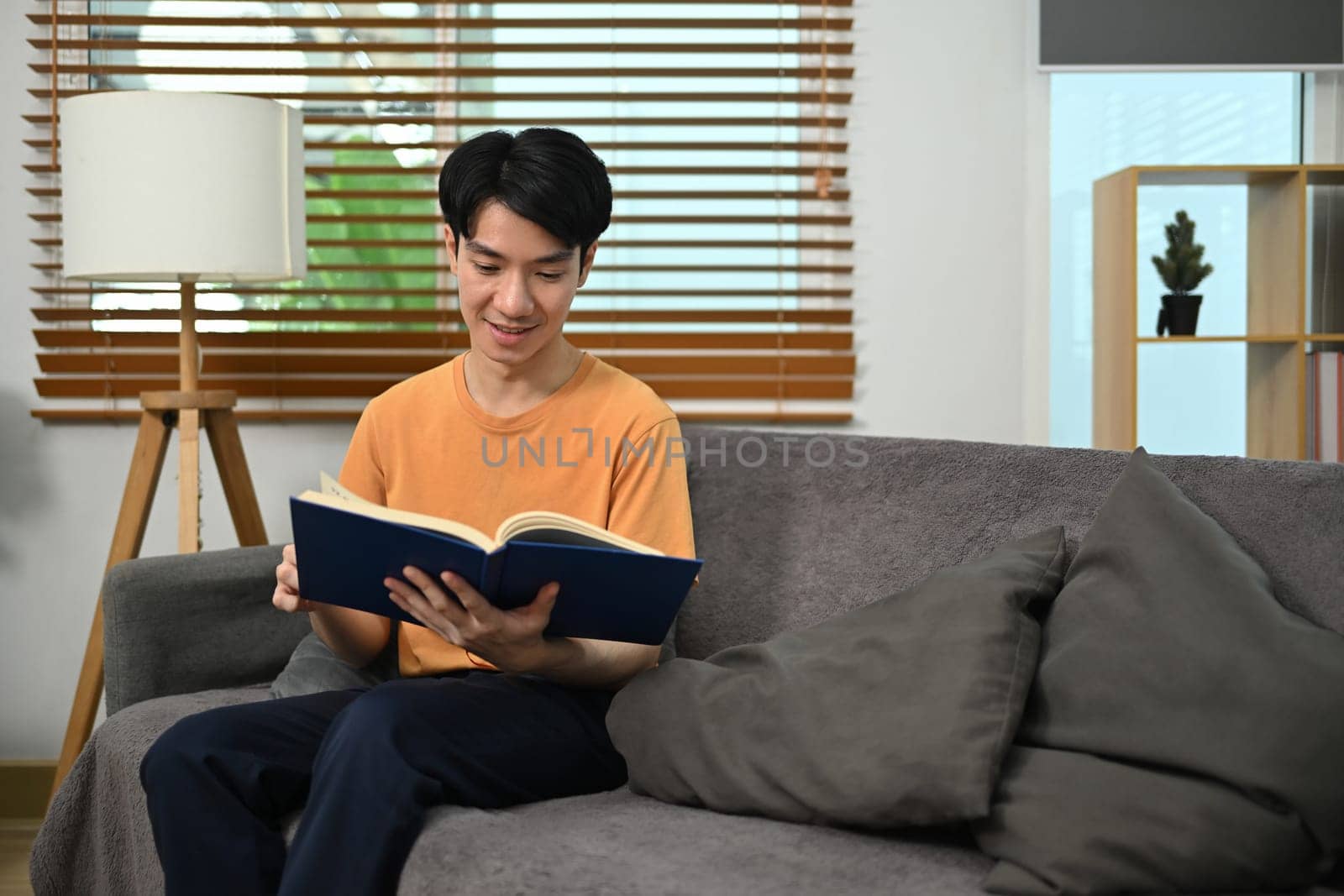 Calm young Asian man in casual clothes reading book in couch at home.