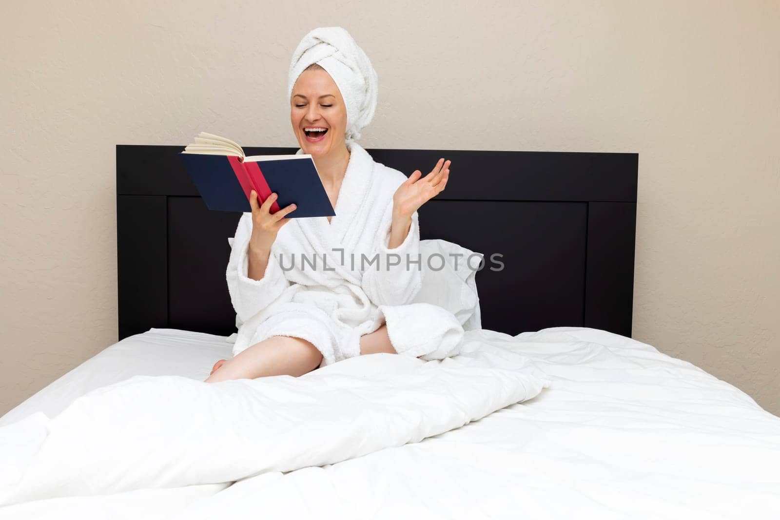 Woman In Her 30s Sits In Bed And Laughing Reading A Book. Emotional Expression Of Face, Smiling, Joy. Caucasian Mature Female Reading Romance, Novel Love Funny Story. Book Lover, Horizontal Plane.