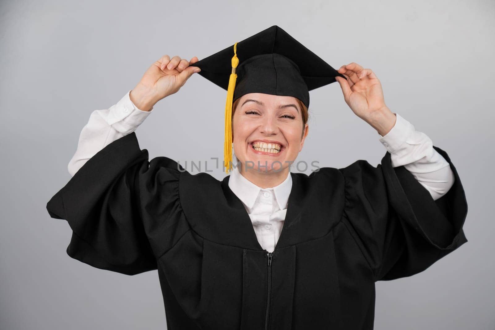 Smiling woman in graduation gown holding cap by tassel on white background