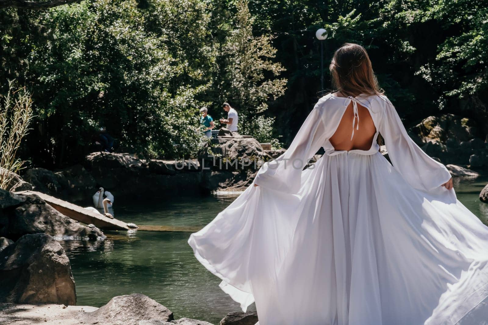 a beautiful woman in a long white dress looks into the distance at a beautiful lake with swans