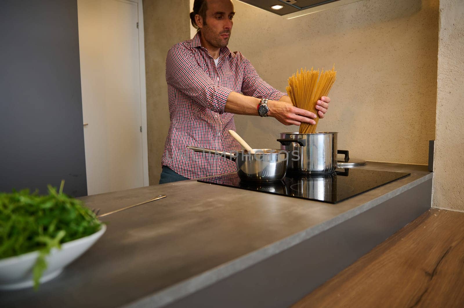 Man chef cook preparing Italian pasta at home. Cropped view of a young man standing at electric stove, putting raw whole grain spaghetti capellini into a stainless steel saucepan with boiling water.