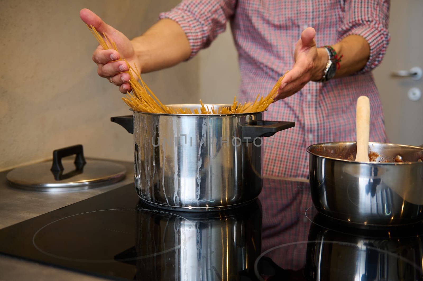 Cropped view of young male chef in plaid shirt, standing at electric stove, putting raw whole grain spaghetti capellini into a stainless steel saucepan with boiling water. Traditional Italian culinary