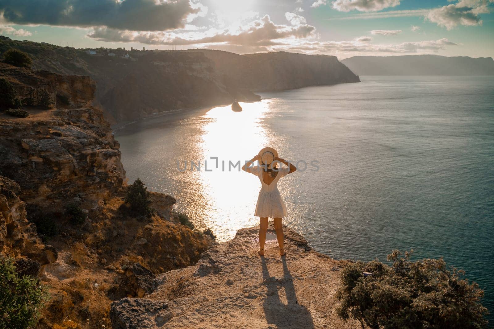 A woman stands on a cliff overlooking the ocean. She is wearing a white dress and a straw hat. The sky is cloudy and the sun is setting