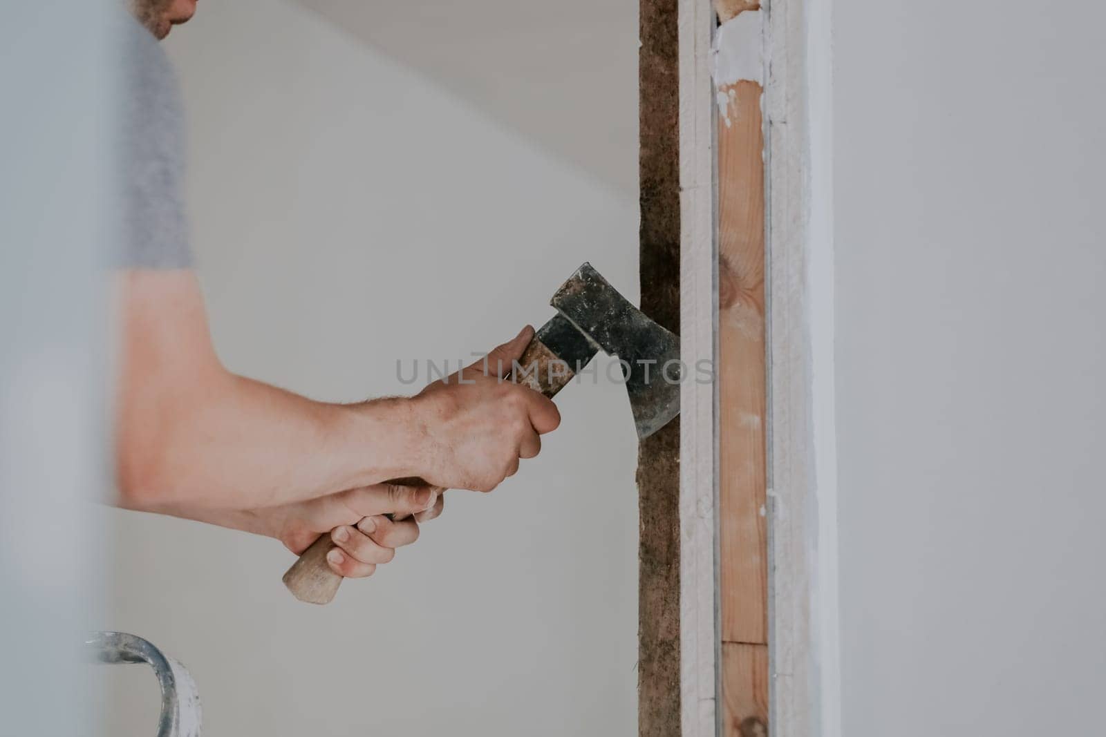 One young unrecognizable Caucasian man in a gray T-shirt is clearing old putty from the beams of a doorway with one hand using an axe, close-up side view with selective focus. Construction work concept.